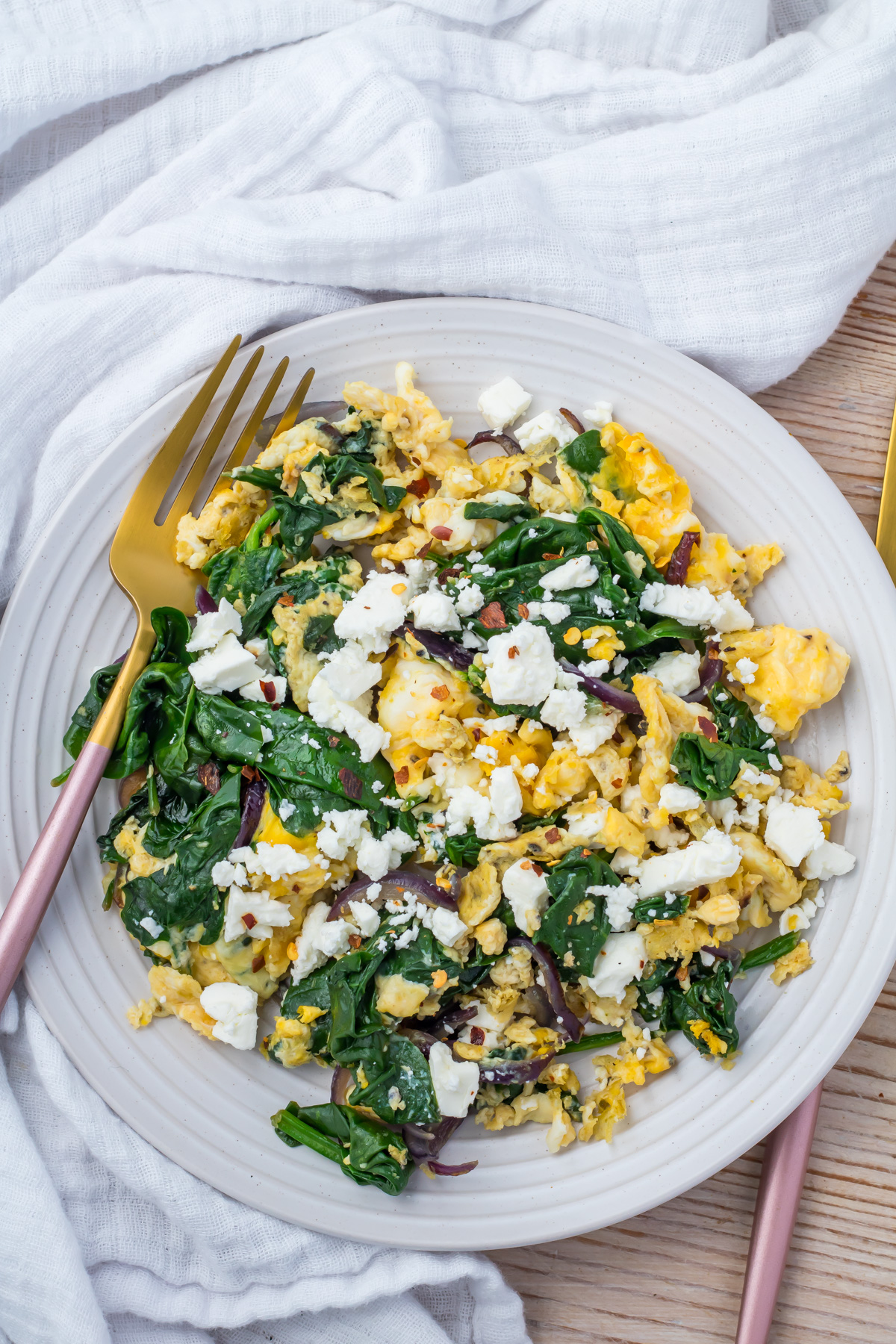 A plate of scrambled eggs with spinach, red onions, and topped with crumbled feta cheese, with a fork placed on the side.