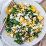 A plate of scrambled eggs with spinach, red onions, and crumbled feta cheese, served with a gold fork on a white plate.