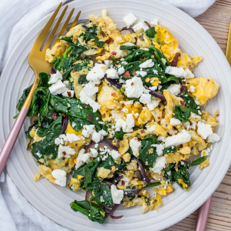 A plate of scrambled eggs with spinach, red onions, and crumbled feta cheese, served with a gold fork on a white plate.