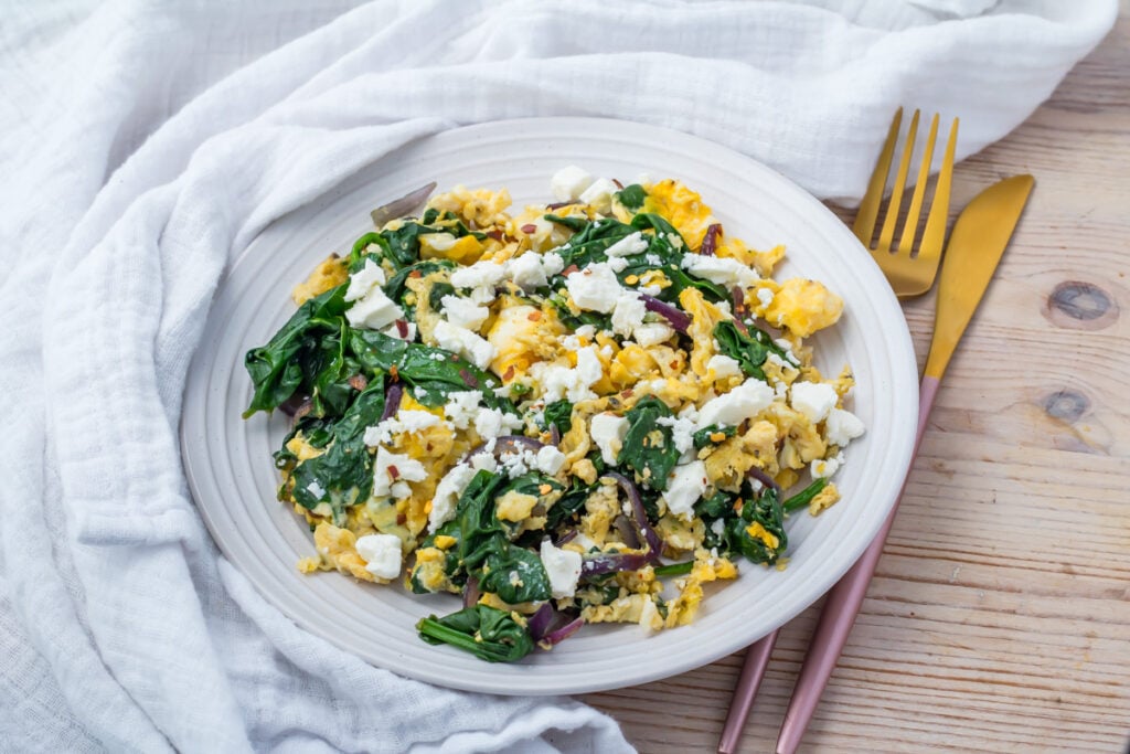 A plate of scrambled eggs with spinach, red onions, and crumbled feta cheese, set on a wooden table with gold and pink cutlery and a white cloth.