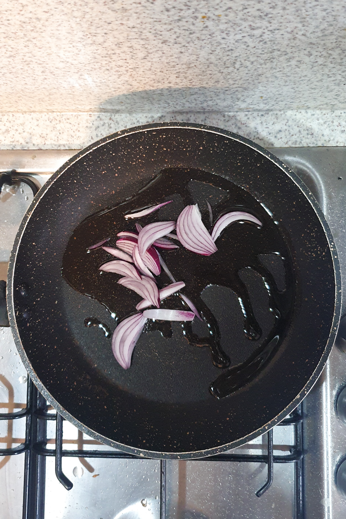 Slices of red onion in a frying pan with oil, placed on a stove with a speckled countertop in the background.