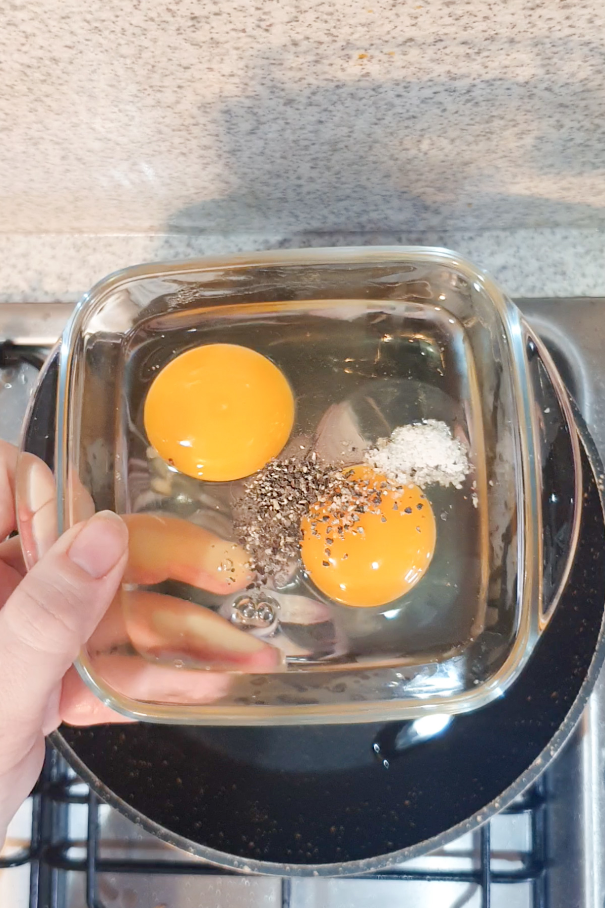 A glass bowl with two raw eggs, salt, and ground black pepper held above a stovetop.