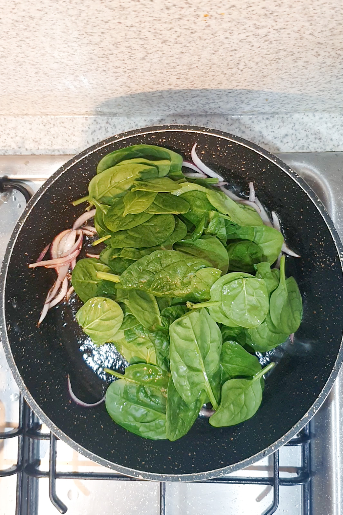 Fresh spinach and sliced red onions cooking in a black frying pan on a gas stove.