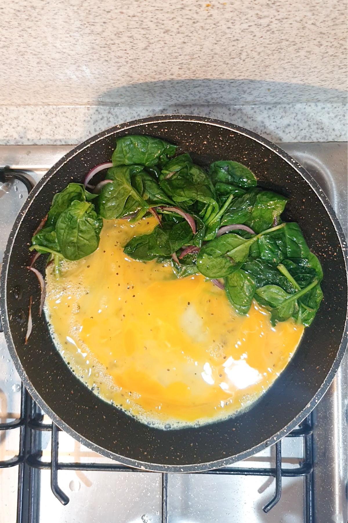 A frying pan on a stove contains beaten eggs and fresh spinach leaves, beginning to cook.