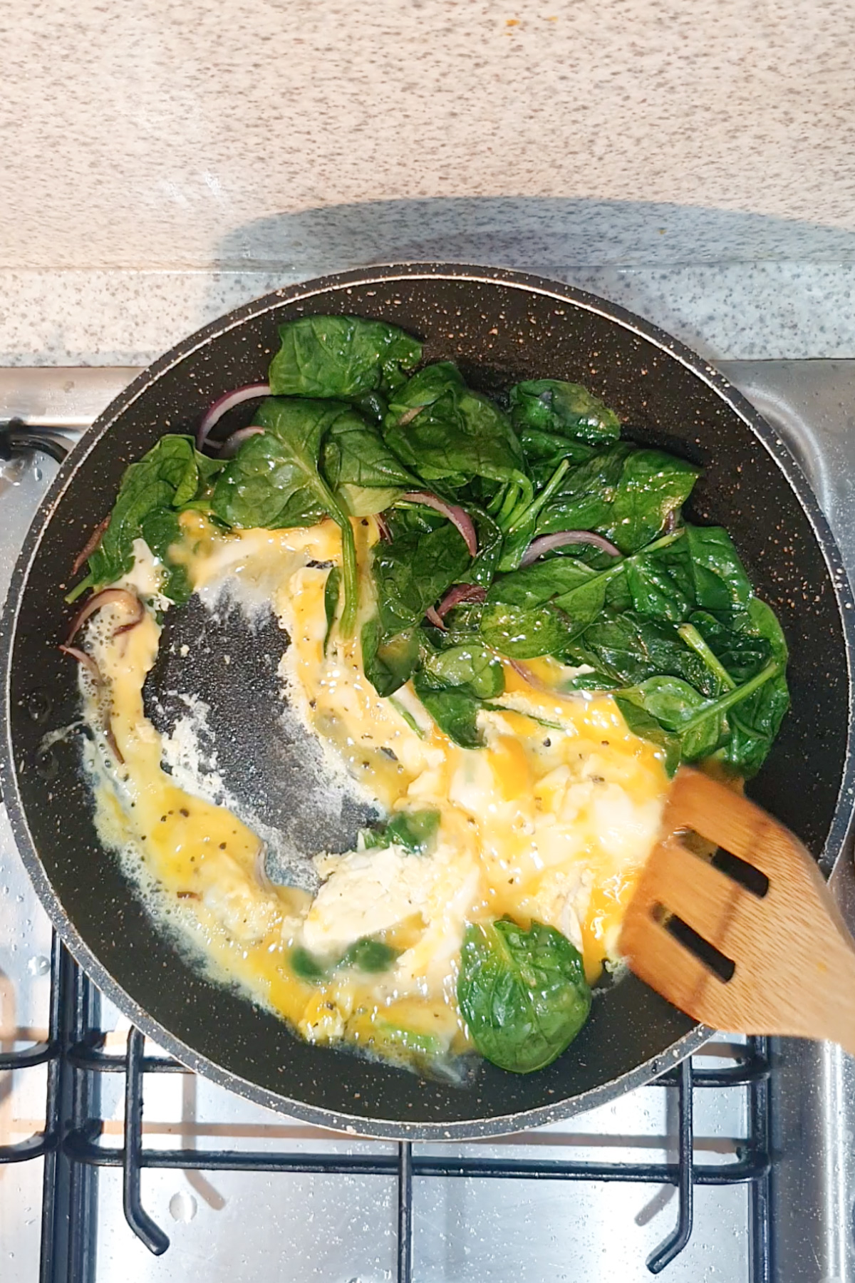 Eggs and fresh spinach being cooked in a nonstick frying pan on a stovetop, stirred with a wooden spatula.