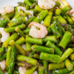 A plate of cooked shrimp and asparagus pieces, seasoned and served with a fork on the side.