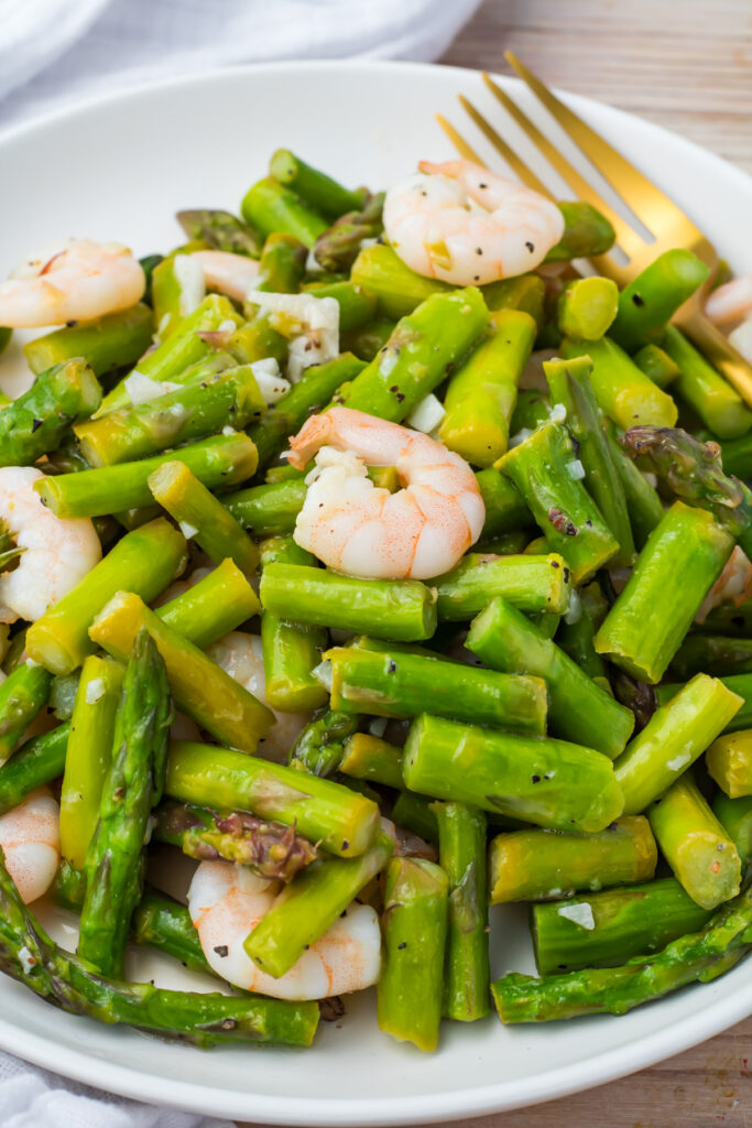 A plate of cooked shrimp and asparagus pieces, seasoned and served with a fork on the side.
