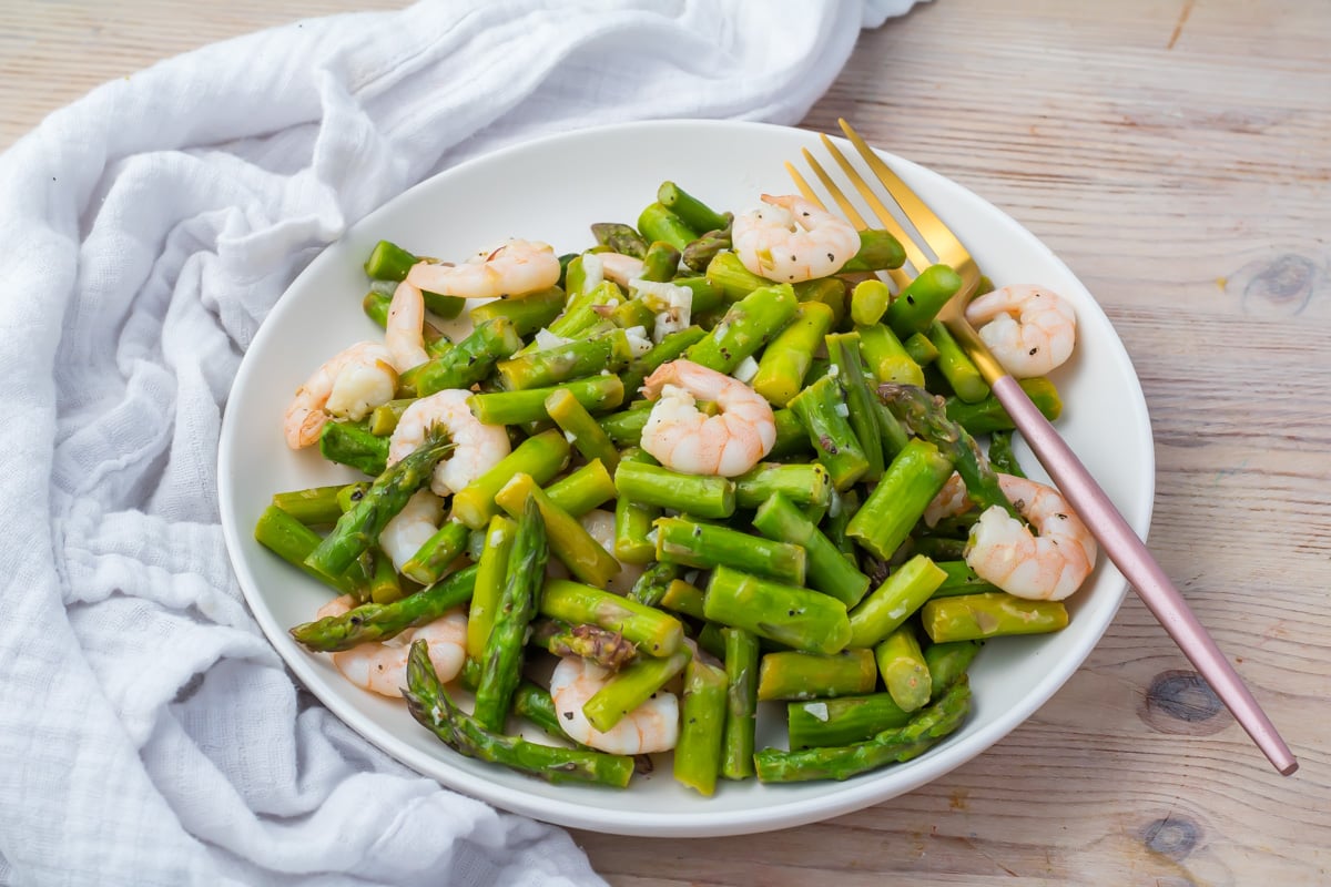 A white plate with cooked shrimp and chopped asparagus, served with a gold and pink fork on a wooden table with a white cloth.
