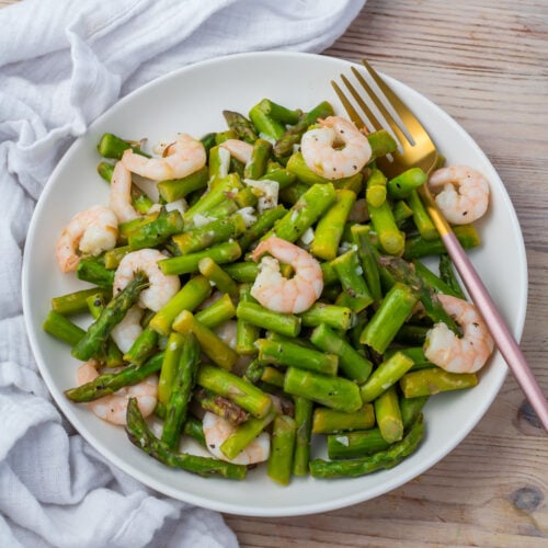 A white plate with cooked shrimp and asparagus, served with a fork on a wooden table with a white cloth nearby.