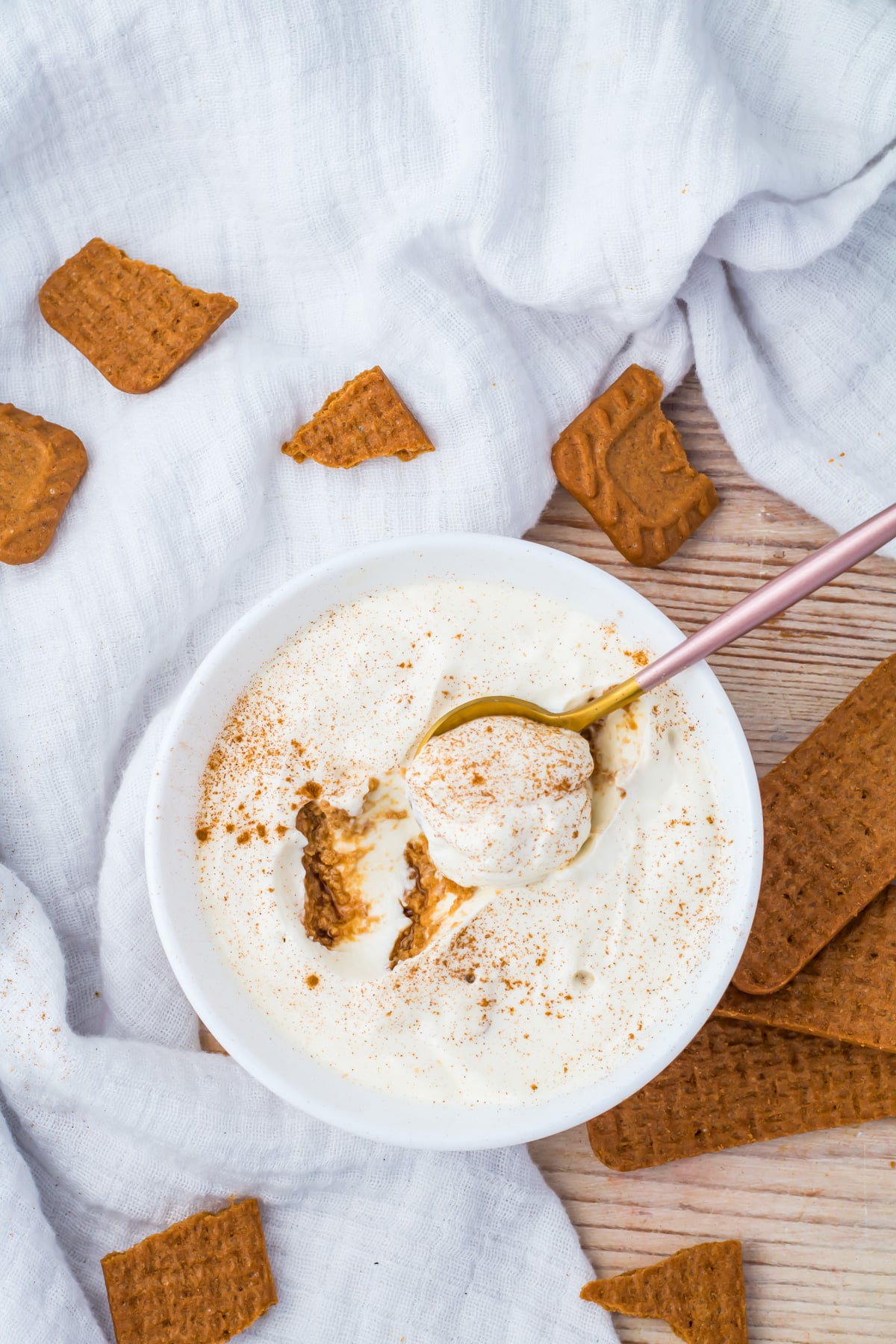 A bowl of 3 ingredient yogurt cheesecake with a spoon, surrounded by pieces of brown cookies on a white cloth.