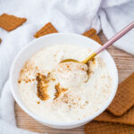 A bowl of 3 ingredient yogurt cheesecake sprinkled with cinnamon, with a spoon scooping some out, surrounded by rectangular brown cookies on a wooden surface.