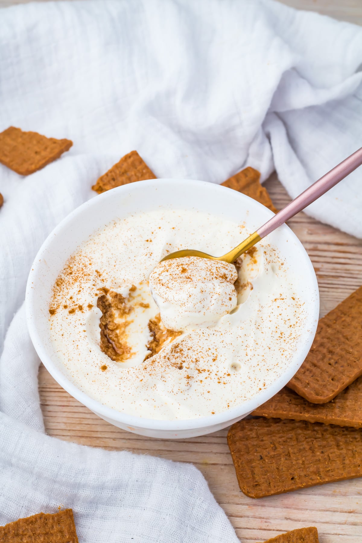 A bowl of 3 ingredient yogurt cheesecake sprinkled with cinnamon, with a spoon scooping some out, surrounded by rectangular brown cookies on a wooden surface.