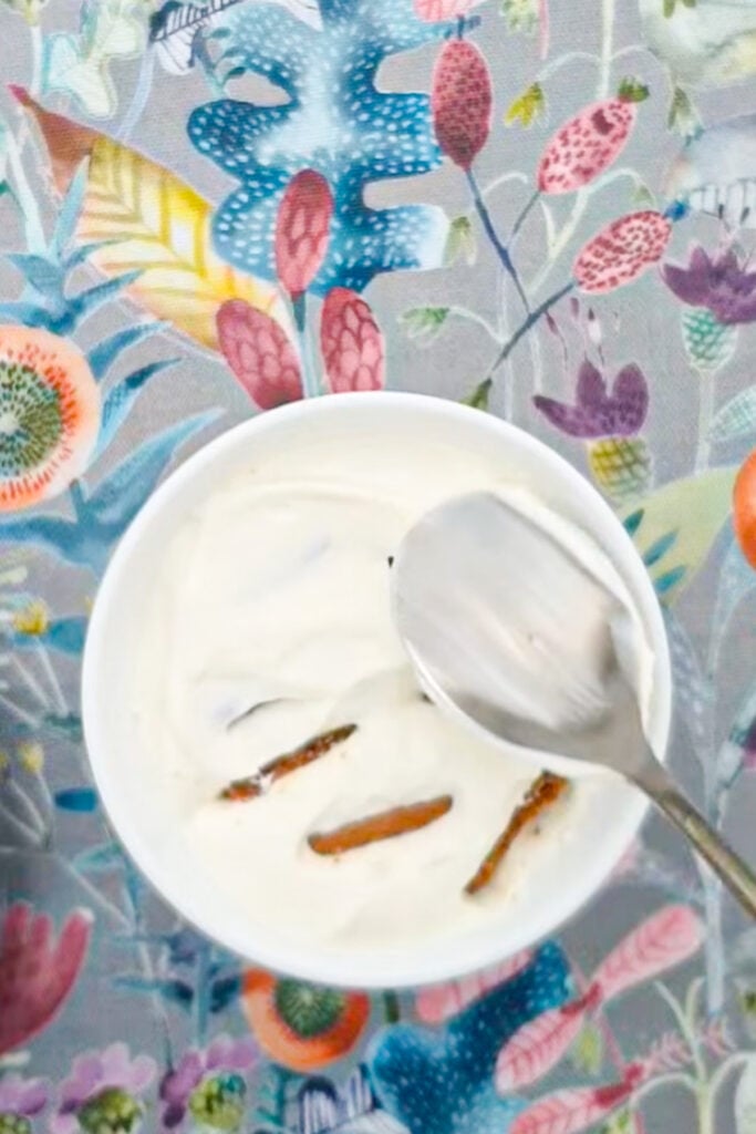 A bowl of white yogurt with pieces of cookies and a spoon sits atop a colorful floral-patterned tablecloth.