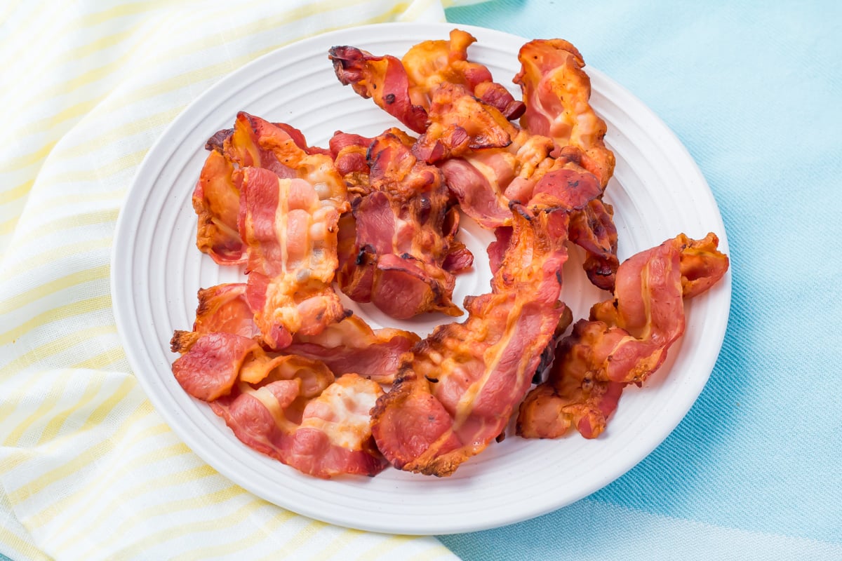 A white plate with several strips of cooked crispy air fryer bacon, placed on a light blue surface with a yellow and white striped cloth nearby.