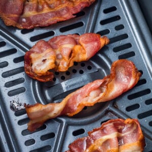 Cooked bacon strips resting on a metal air fryer tray with visible grooves and a few spots of grease.