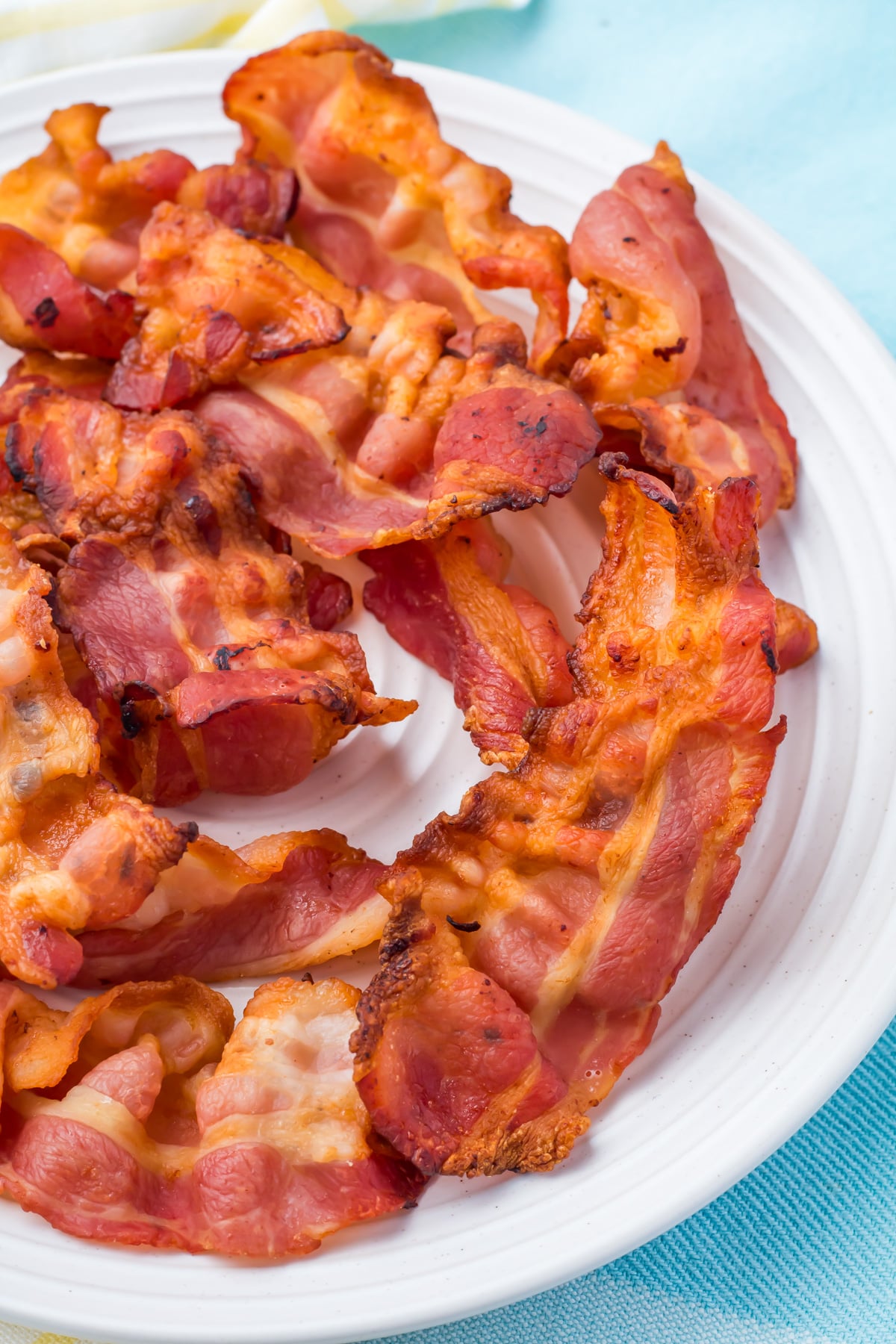 A white plate filled with several strips of cooked crispy air fryer bacon, set on a light blue and yellow background.