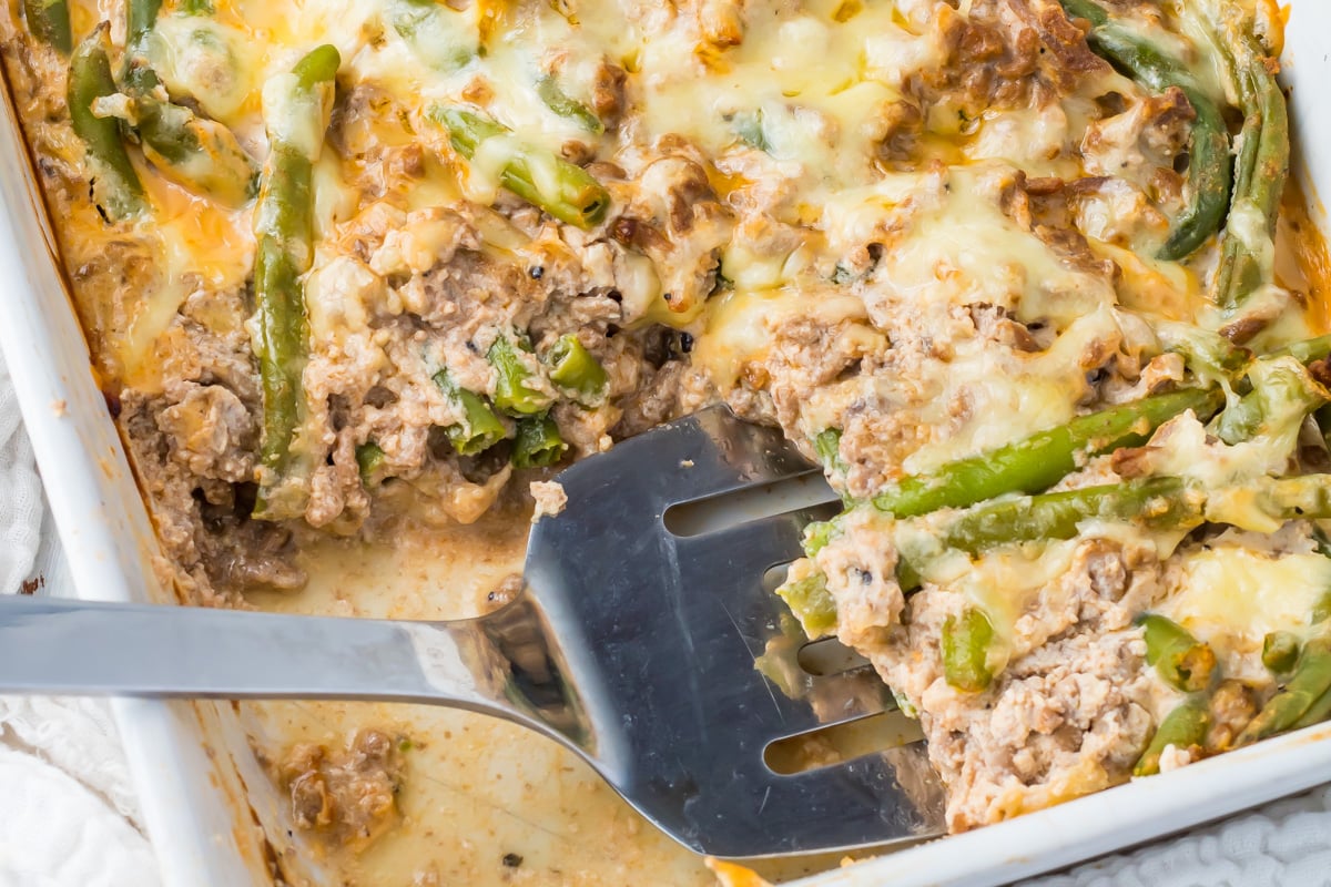 Keto green bean hamburger casserole with ground meat, green beans, and melted cheese, partially served with a metal spatula in a white baking dish.