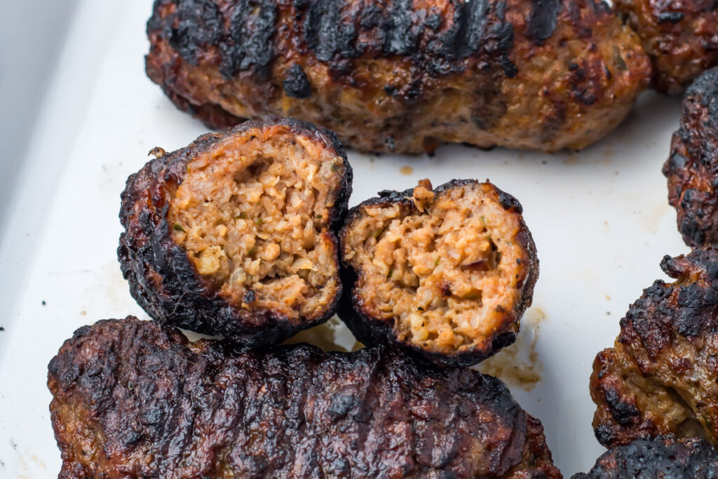 Grilled lamb kofta kebabs, one split open to show the cooked, seasoned interior, arranged on a white surface.