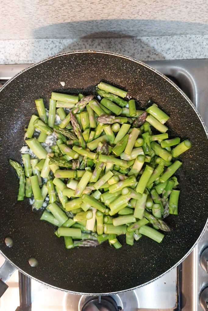 Chopped asparagus pieces and tender shrimp being sautéed in a black nonstick pan on a gas stove.