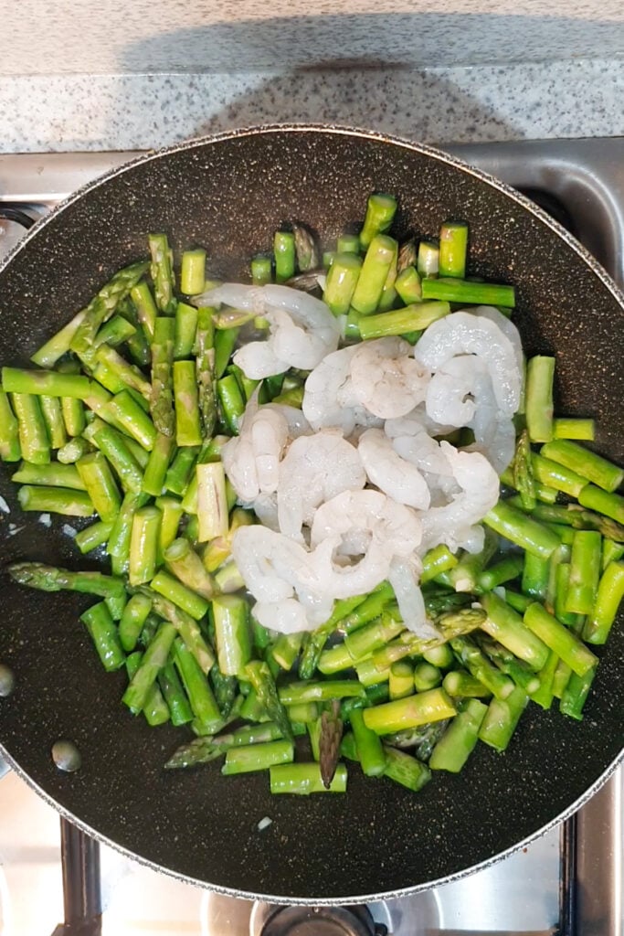 Shrimp and asparagus are being cooked together in a frying pan on a stovetop, with the chopped asparagus and raw shrimp sizzling side by side.