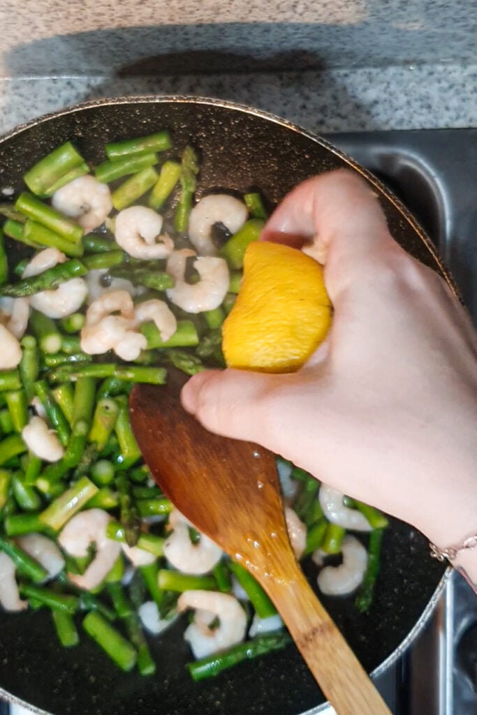 A hand squeezes lemon over shrimp and asparagus cooking in a pan, with a wooden spoon stirring the vibrant shrimp and asparagus mixture on a stovetop.