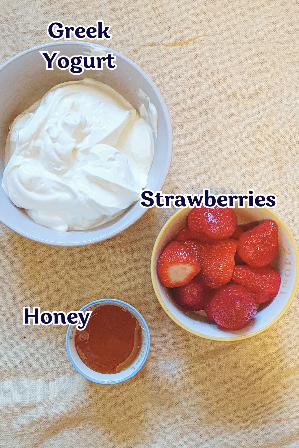 A bowl of Greek yogurt, a bowl of strawberries, and a small dish of honey are arranged on a light-colored cloth. Each item is labeled with text.