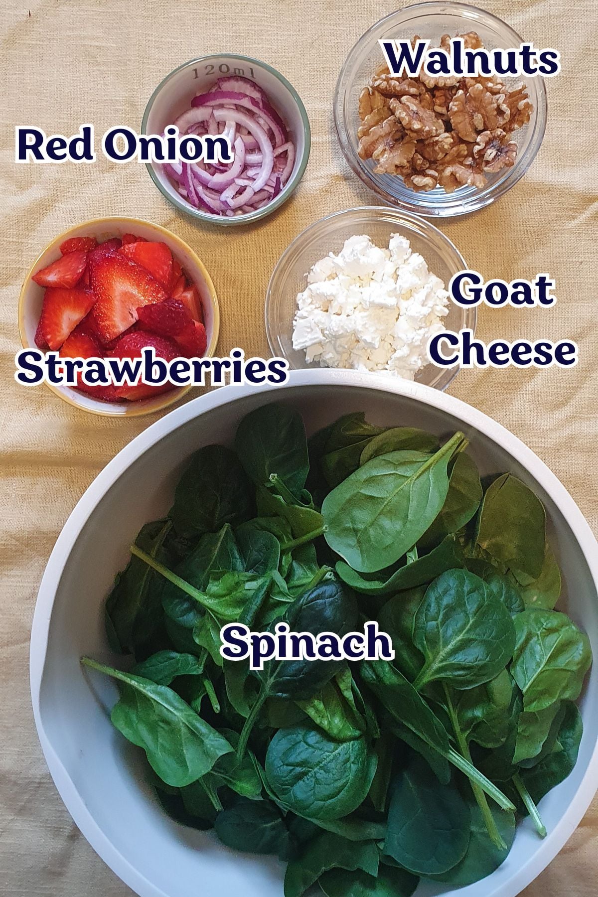 Bowls containing spinach, sliced strawberries, sliced red onion, walnuts, and crumbled goat cheese are arranged on a light surface.