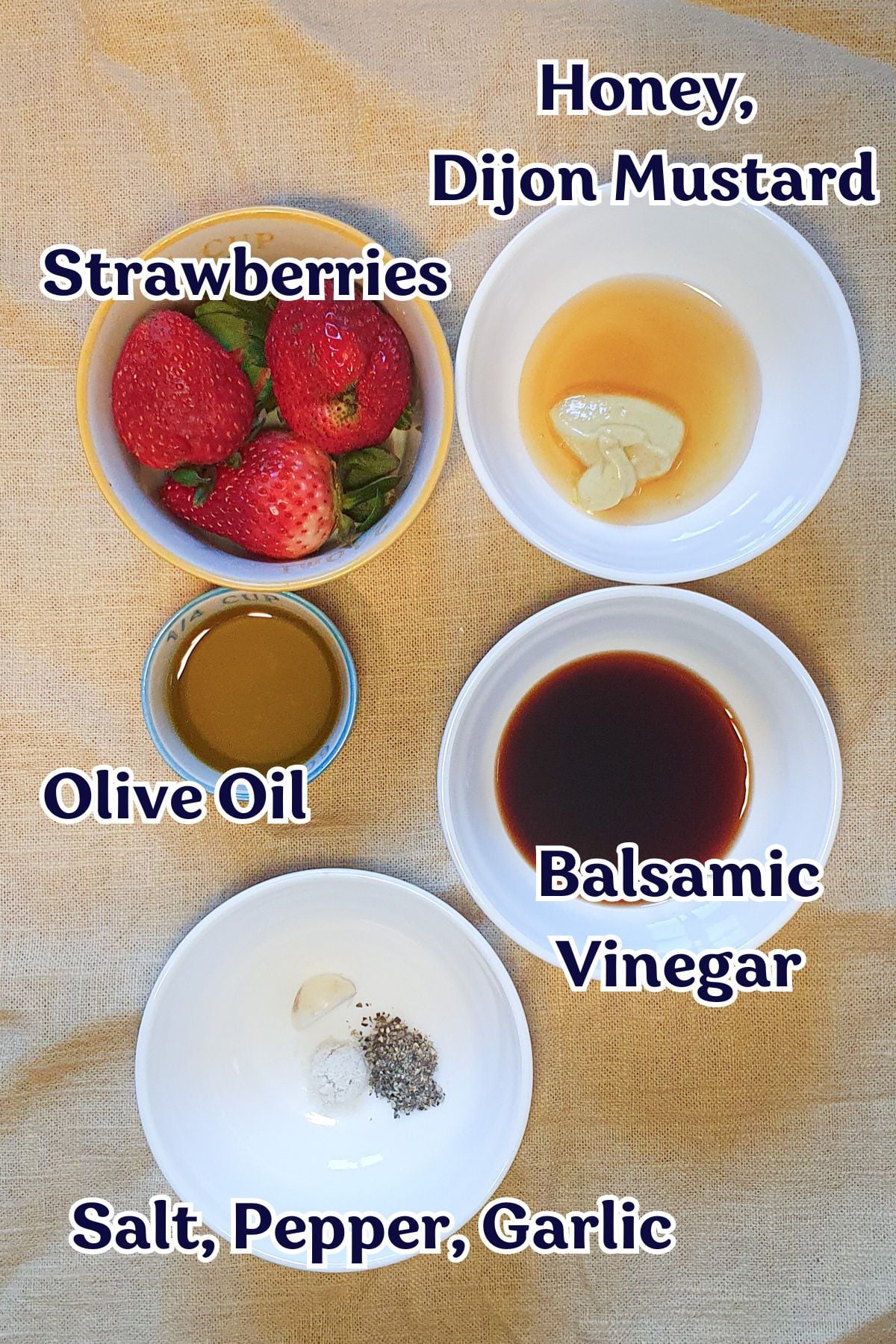 Five bowls containing strawberries, olive oil, honey with Dijon mustard, balsamic vinegar, and a mix of salt, pepper, and garlic arranged on a beige cloth.