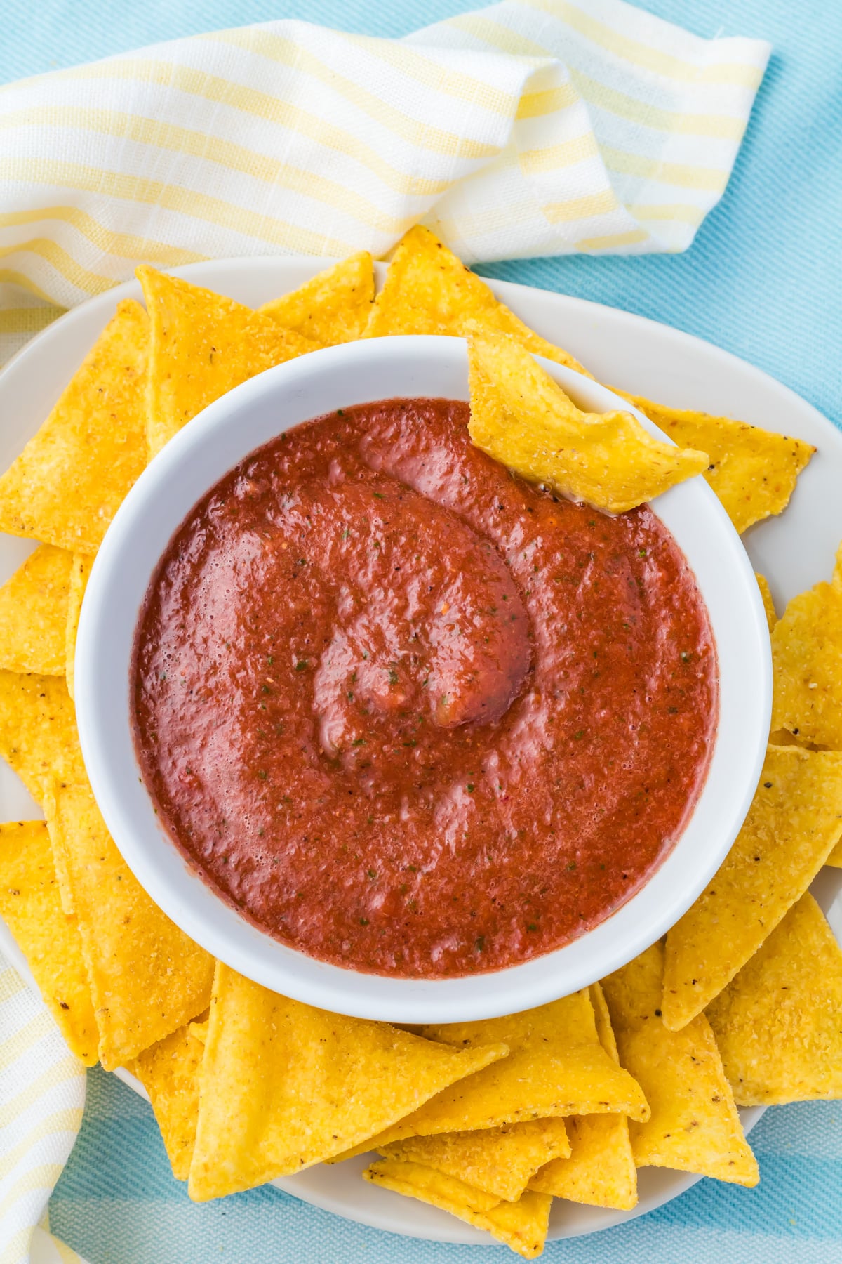 A white bowl of red blender salsa surrounded by yellow tortilla chips on a white plate, with a yellow and white striped cloth in the background.