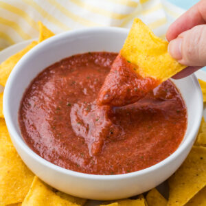 A hand dips a tortilla chip into a bowl of red blender salsa, surrounded by more chips.