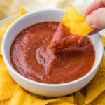 A hand dips a tortilla chip into a bowl of red blender salsa, surrounded by more tortilla chips on a plate.