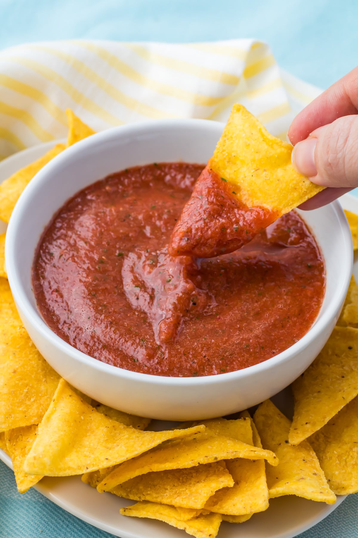 A hand dips a tortilla chip into a bowl of red blender salsa, surrounded by more tortilla chips on a plate.