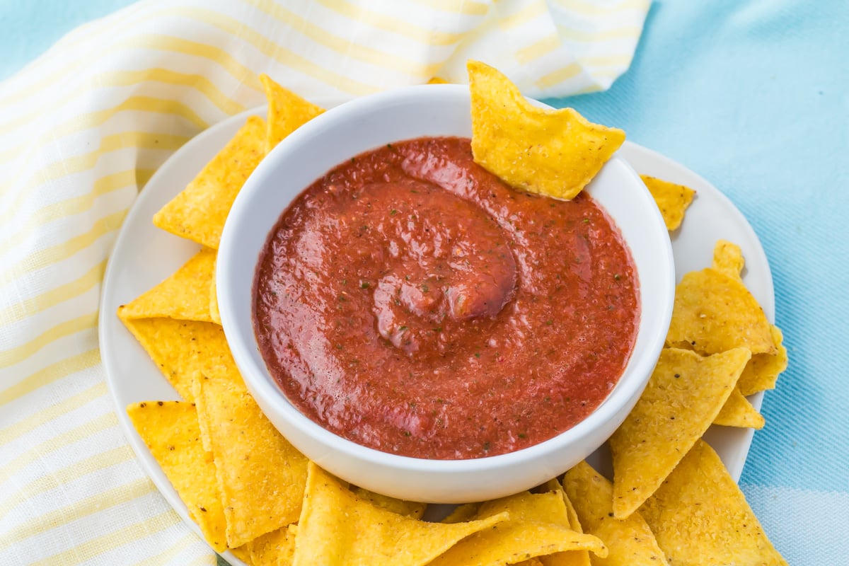 A white bowl of red blender salsa is surrounded by yellow tortilla chips on a white plate, with a striped cloth and blue background.