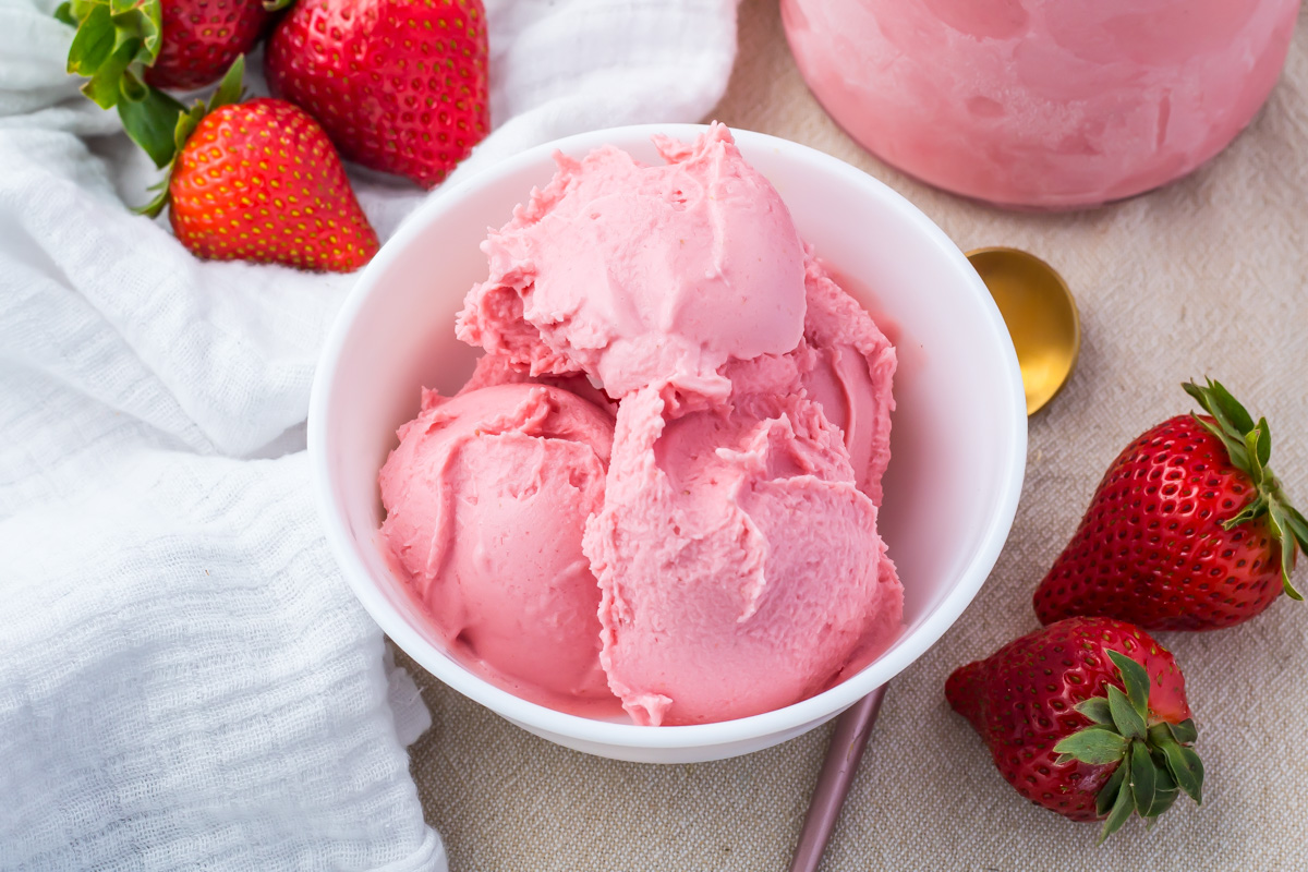 A white bowl filled with scoops of Ninja Creami Strawberry Frozen Yogurt, surrounded by fresh strawberries on a beige surface with a gold spoon nearby.