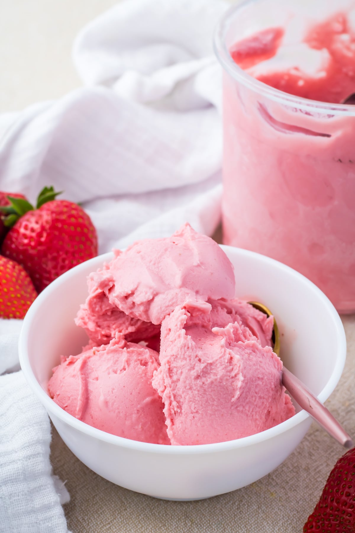 A white bowl filled with scoops of Ninja Creami Strawberry Frozen Yogurt sits next to fresh strawberries and a jar of more ice cream.