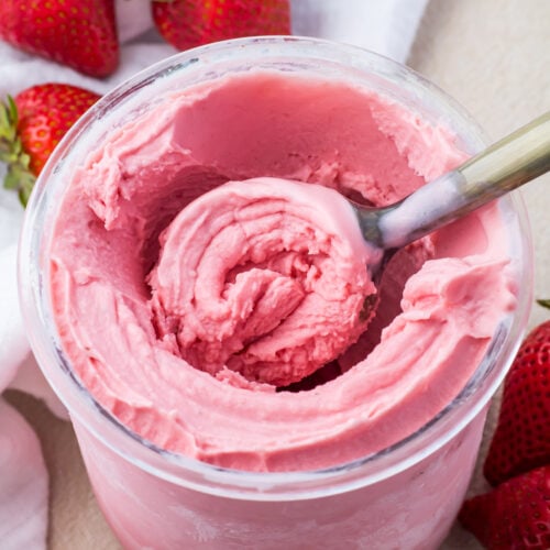 A scoop is dipped into a jar of Ninja Creami Strawberry Frozen Yogurt, surrounded by fresh strawberries.