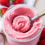 A scoop in a jar of Ninja Creami Strawberry Frozen Yogurt with fresh strawberries and a white cloth nearby.