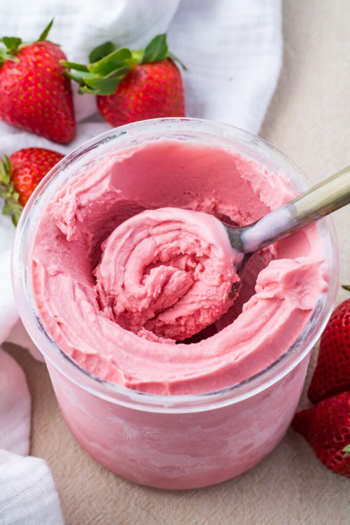 A scoop in a jar of Ninja Creami Strawberry Frozen Yogurt with fresh strawberries and a white cloth nearby.