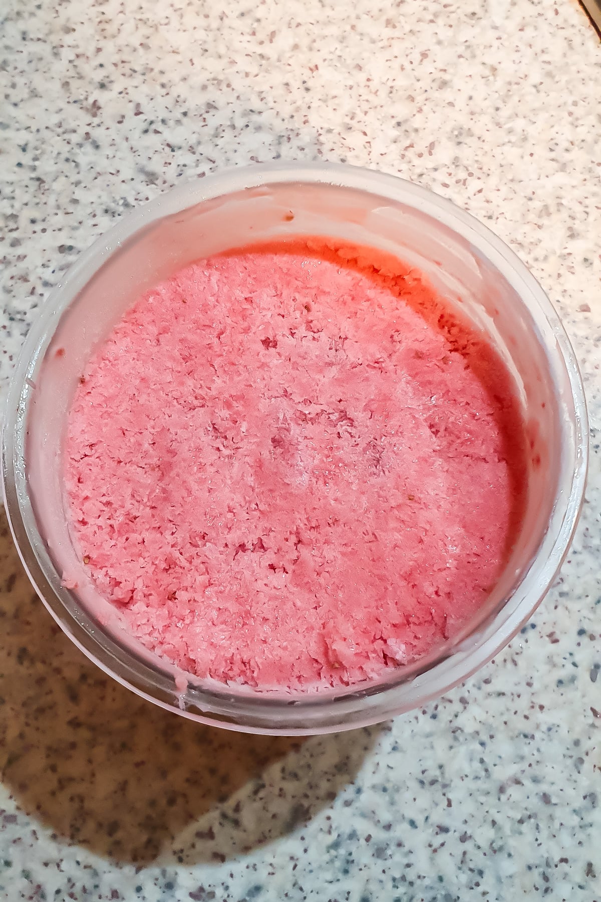 A round plastic container filled with a pink, textured frozen substance sits on a speckled countertop.