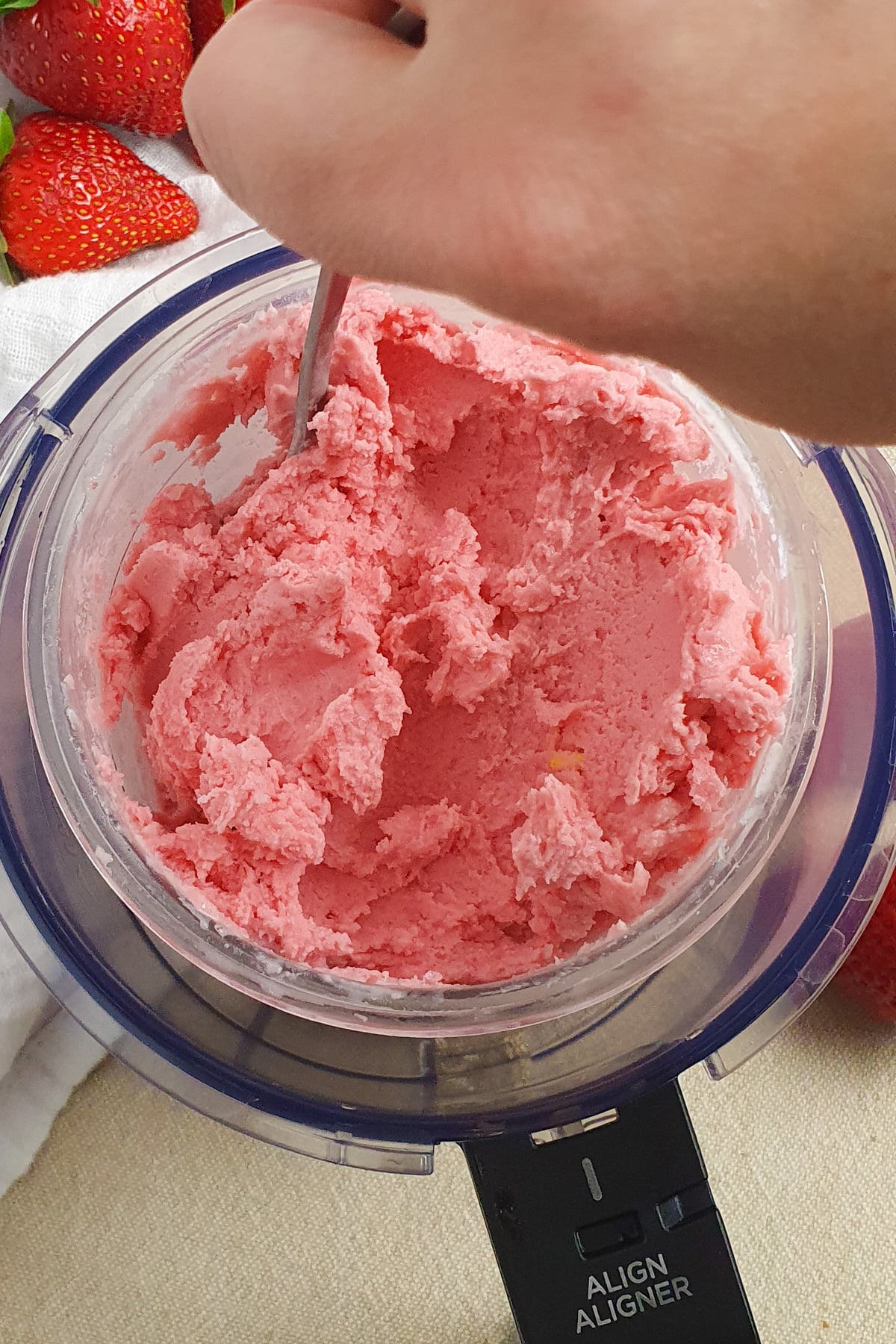 A hand holds a spoon, scooping pink Ninja Creami Strawberry Frozen Yogurt from the sides of the pint, with fresh strawberries nearby.