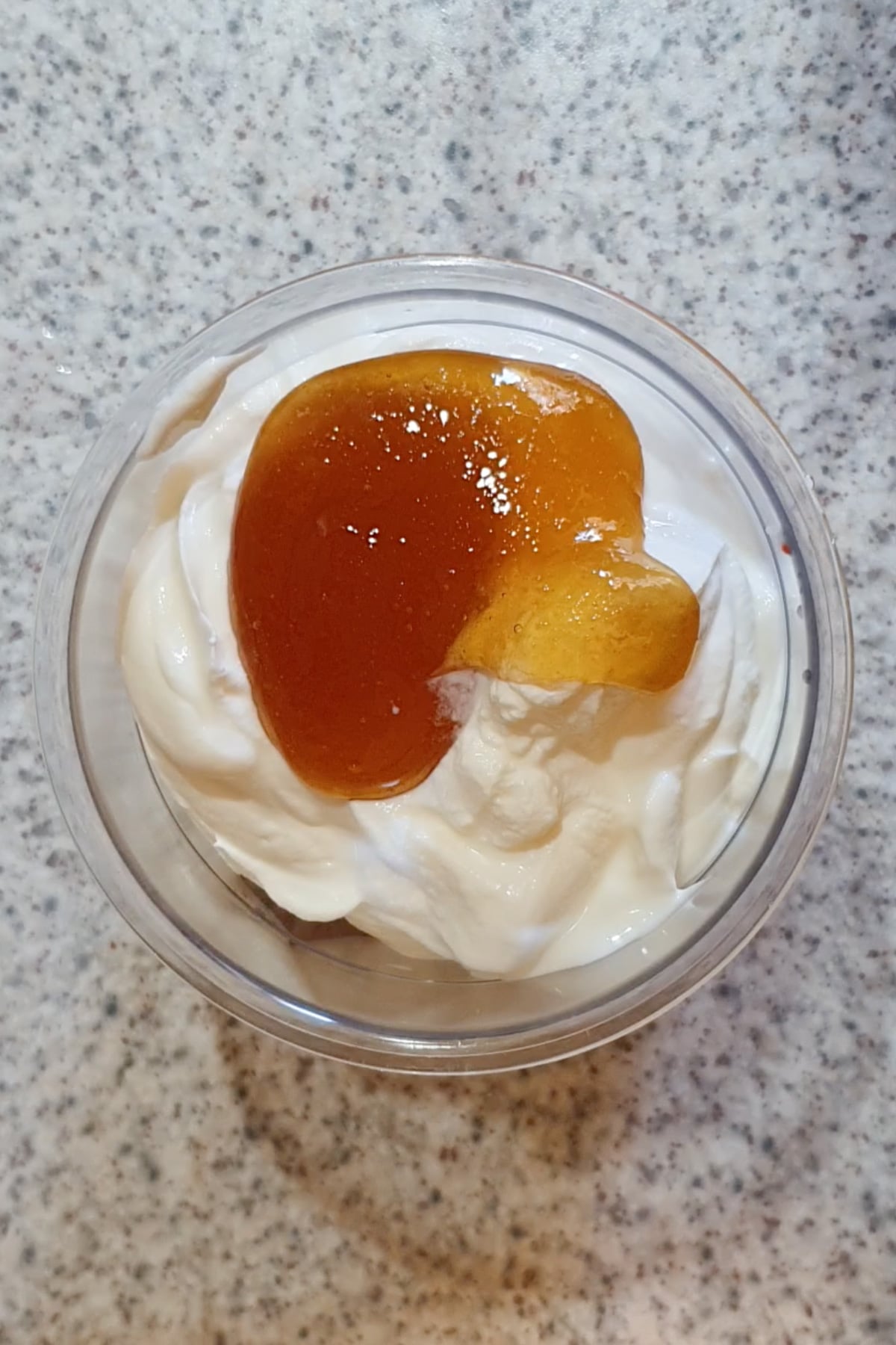 A clear cup filled with white yogurt topped with a dollop of golden honey, placed on a speckled countertop.