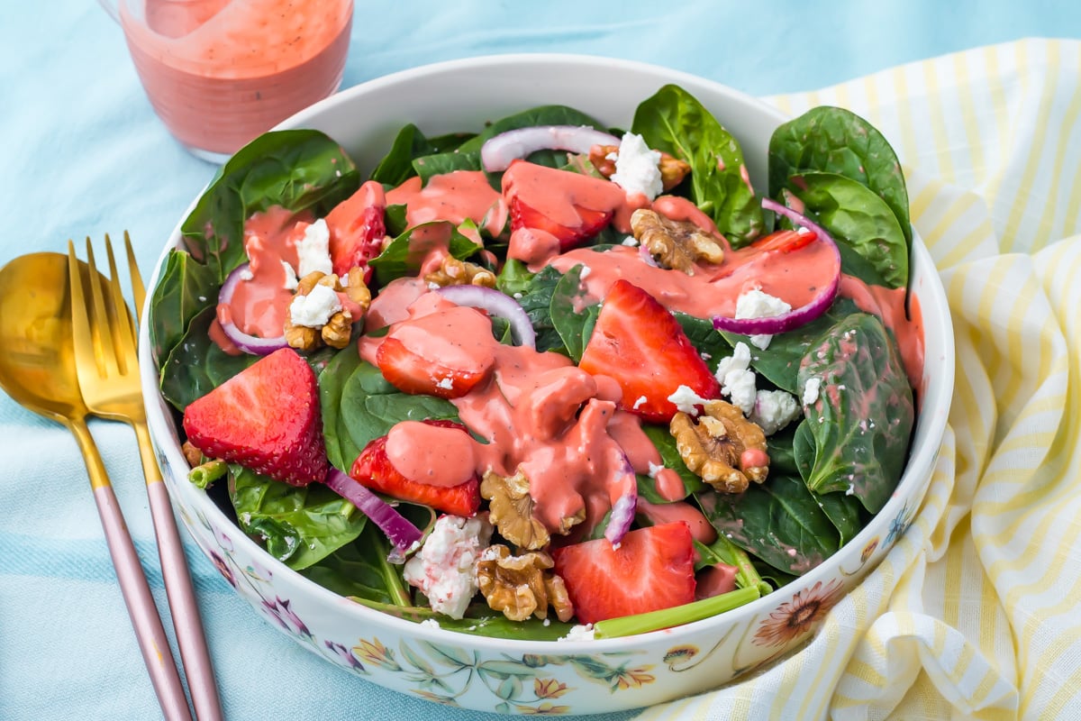 A bowl of strawberry salad with goat cheese, with spinach, strawberries, walnuts, red onion, goat cheese, and pink dressing, placed beside gold and copper utensils on a blue tablecloth.