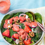 A bowl of strawberry salad with goat cheese, with strawberries, walnuts, red onions, goat cheese, and pink dressing, with a jar of dressing and utensils on the side.