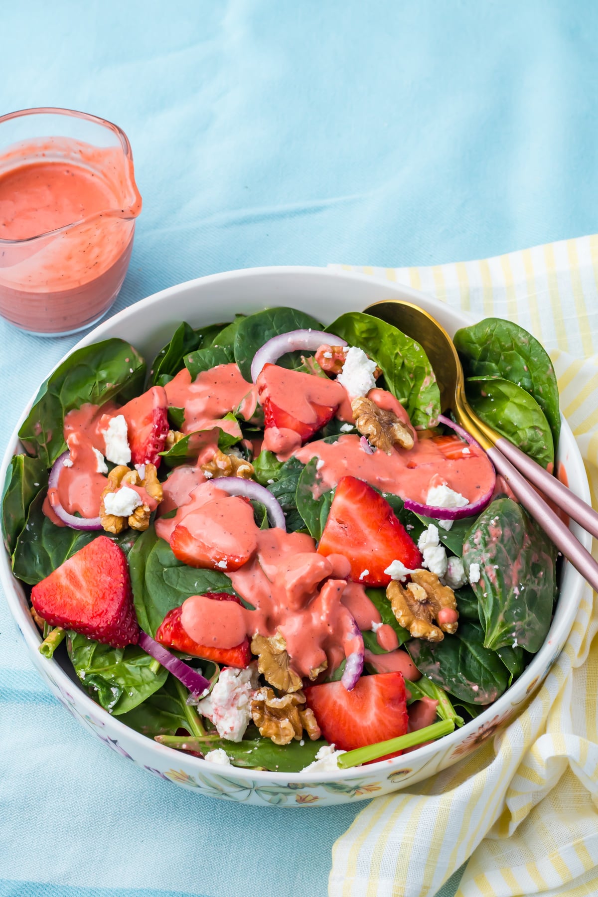 A bowl of strawberry salad with goat cheese, with strawberries, walnuts, red onions, goat cheese, and pink dressing, with a jar of dressing and utensils on the side.