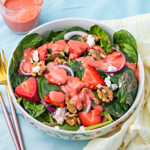 A bowl of strawberry salad with goat cheese, with strawberries, walnuts, red onion, goat cheese, spinach and pink dressing, with a fork and a small pitcher of dressing beside it.