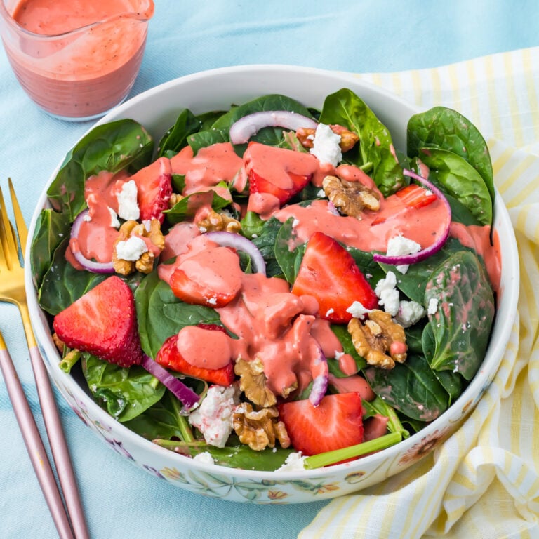A bowl of strawberry salad with goat cheese, with strawberries, walnuts, red onion, goat cheese, spinach and pink dressing, with a fork and a small pitcher of dressing beside it.