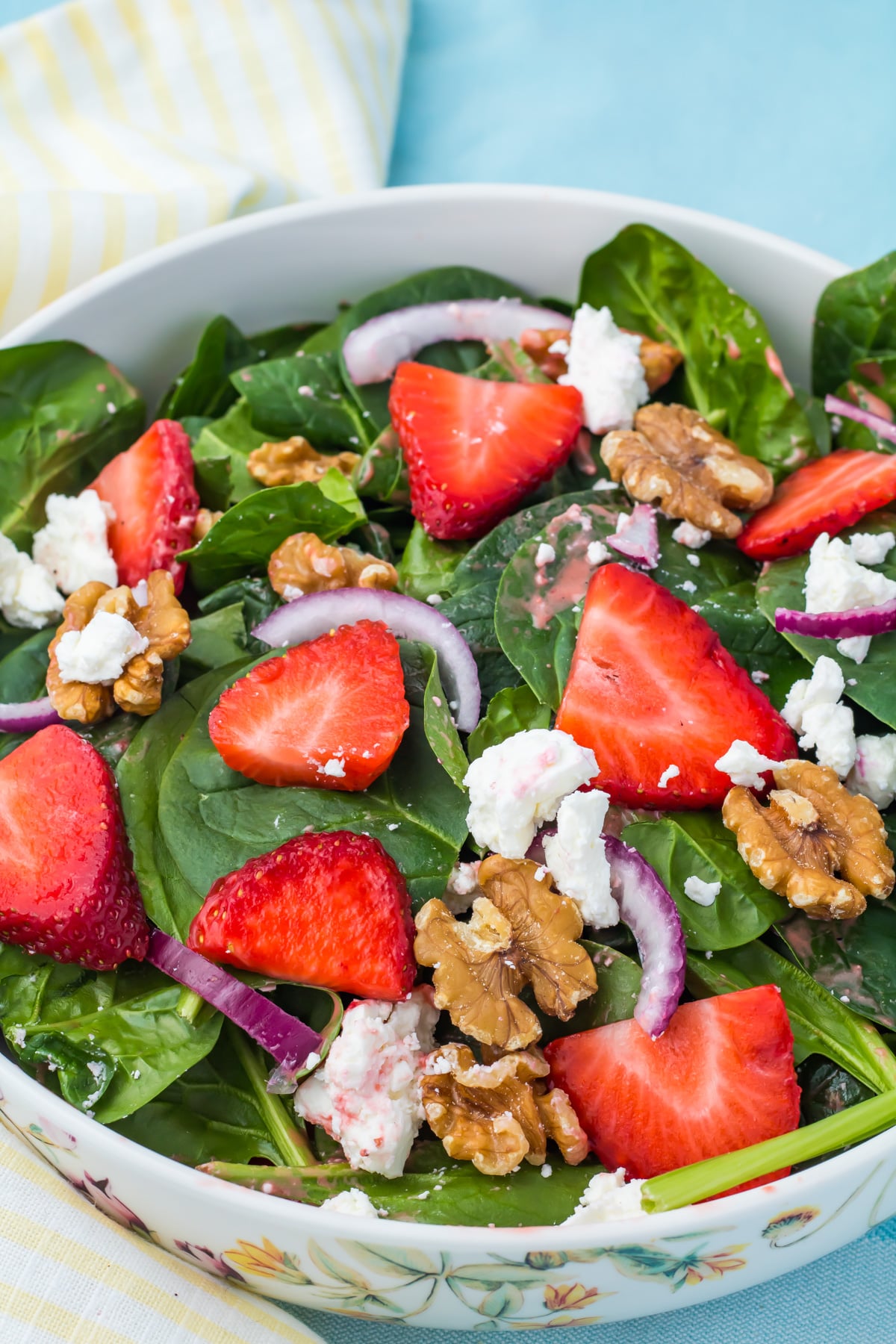 A bowl of strawberry salad with goat cheese, topped with sliced strawberries, walnuts, spinach, red onion, and crumbled cheese on a blue surface with a striped napkin.