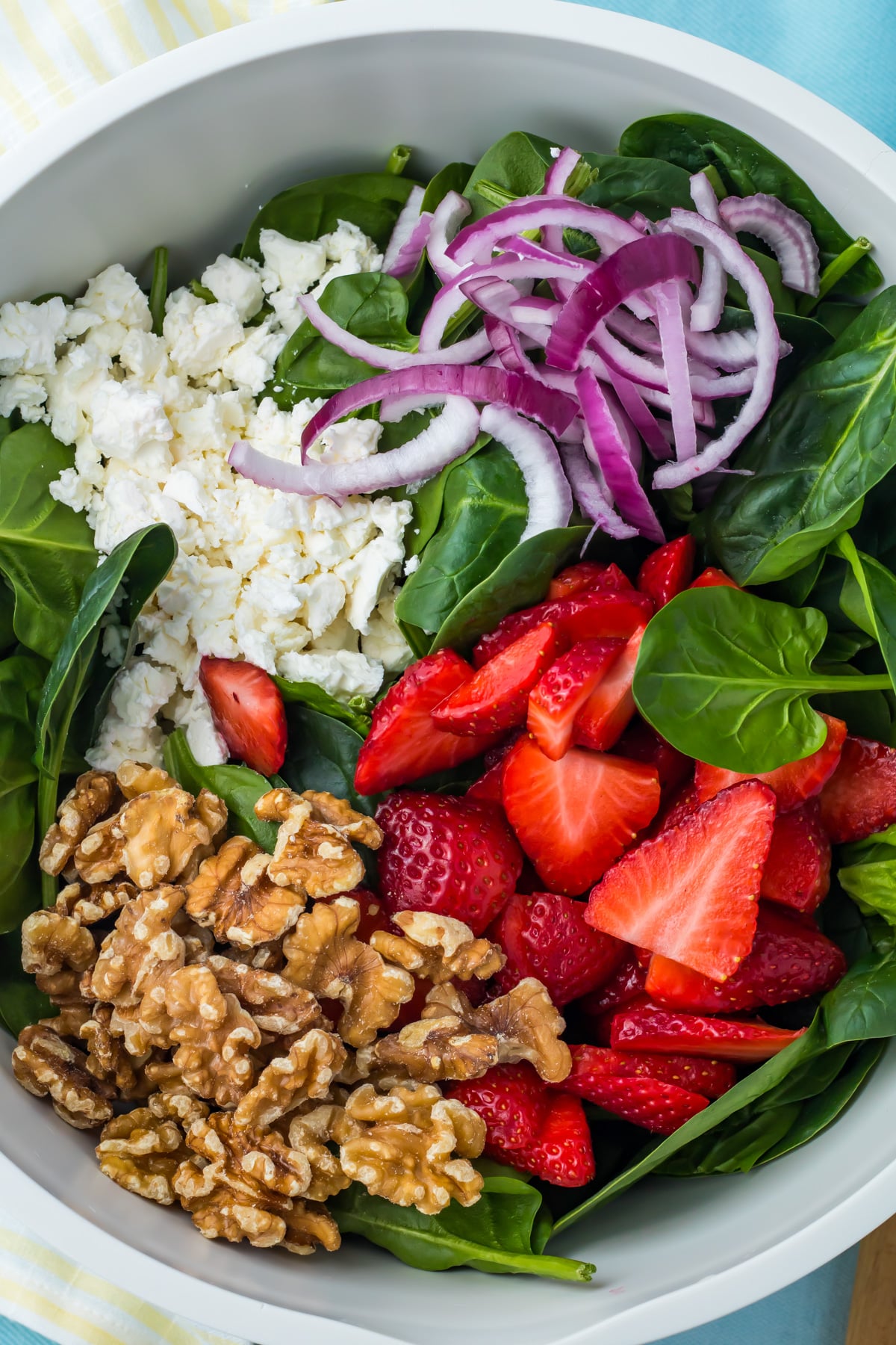 A salad bowl contains fresh spinach leaves, sliced strawberries, walnuts, crumbled goat cheese, and sliced red onions, arranged in separate sections.