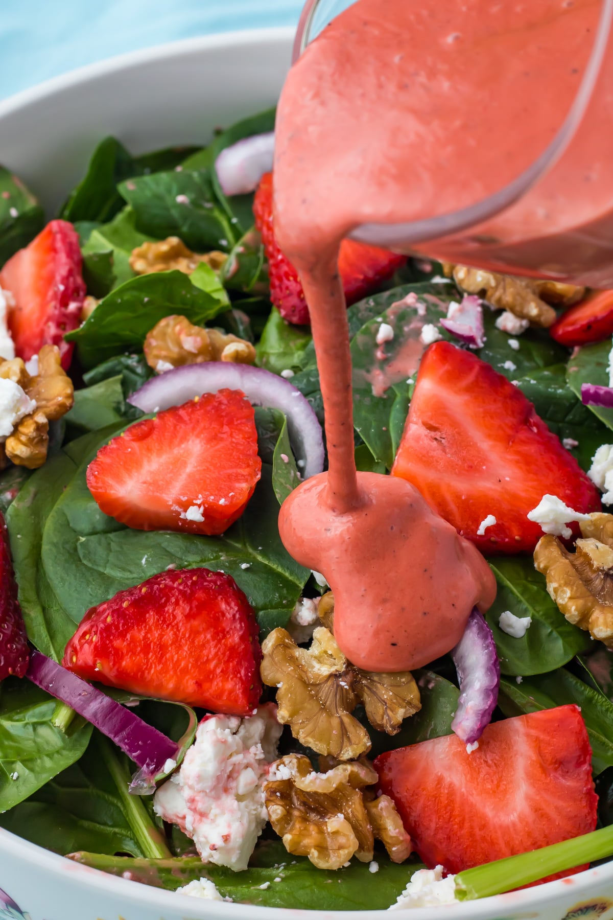 A glass container pours pink strawberry vinaigrette over a salad with spinach, strawberries, walnuts, red onion, and feta cheese.
