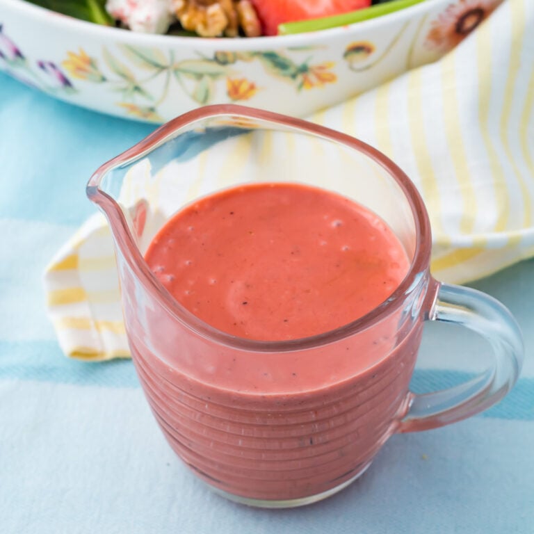 A glass pitcher filled with strawberry vinaigrette sits on a table next to a striped yellow napkin and a bowl of salad.